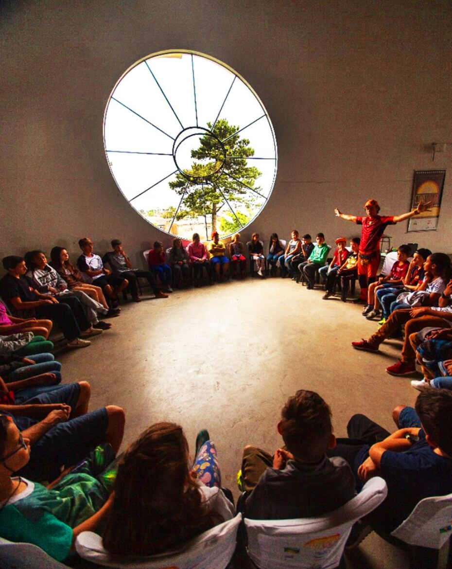 Grande grupo de crianças e jovens sentados em círculo dentro de uma sala ampla, com um orientador em pé à direita gesticulando, sob a iluminação natural vinda de uma grande janela circular