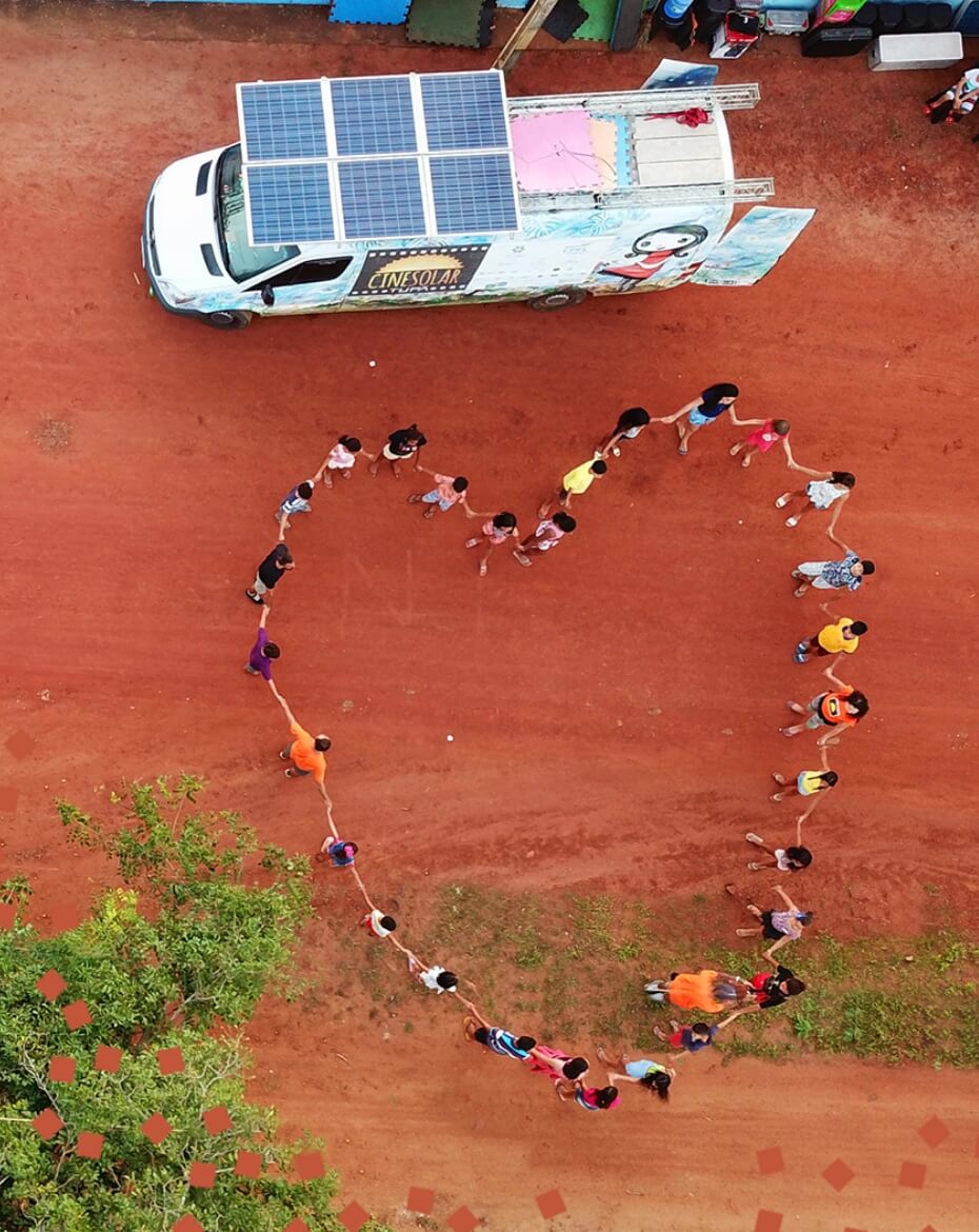 Vista aérea de um grupo de pessoas de mãos dadas formando o desenho de um grande coração em um chão de terra vermelha, posicionado logo abaixo do furgão da CineSolar