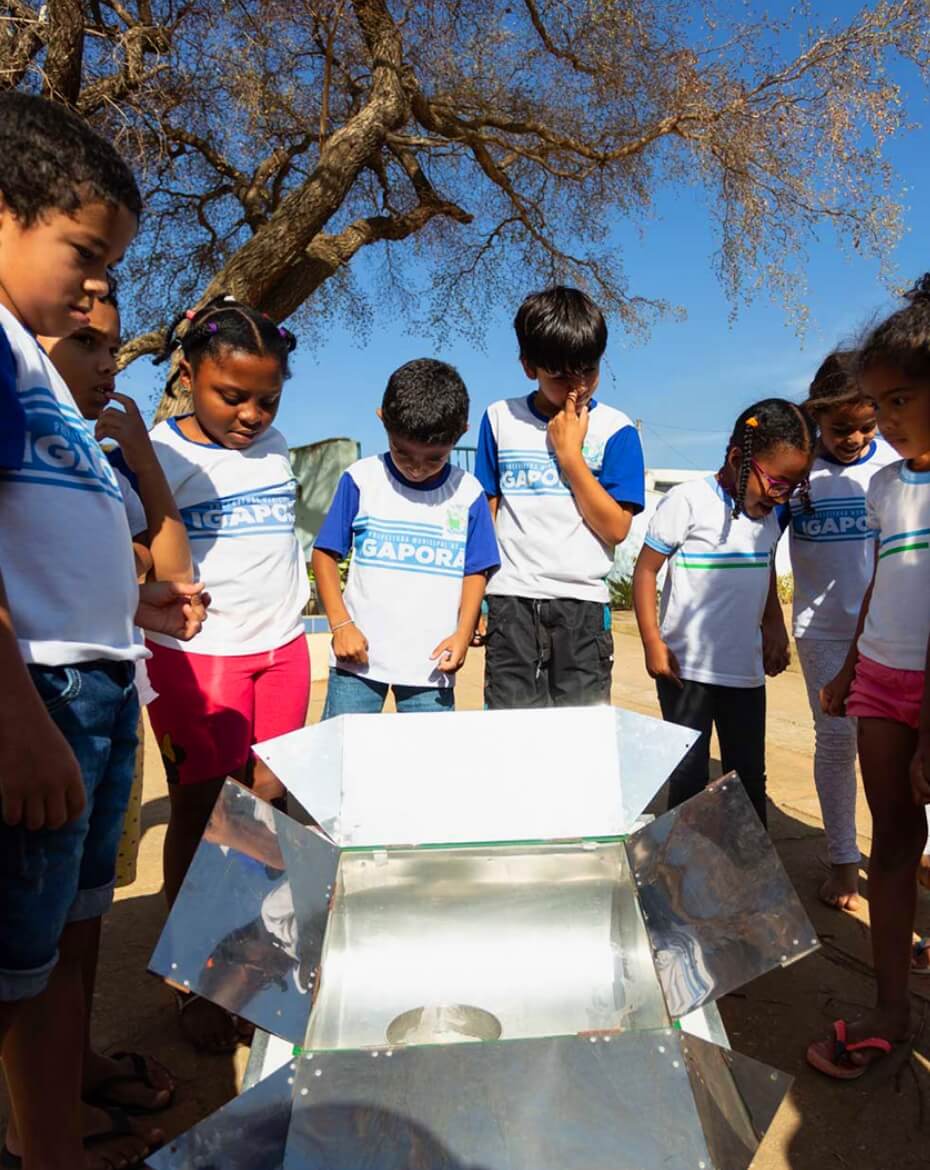 Grupo de crianças vestindo uniformes escolares reunido ao ar livre sob o sol, observando curiosamente o interior de um forno solar portátil com painéis refletores metálicos aberto à sua frente, tendo uma árvore de galhos secos e o céu azul ao fundo