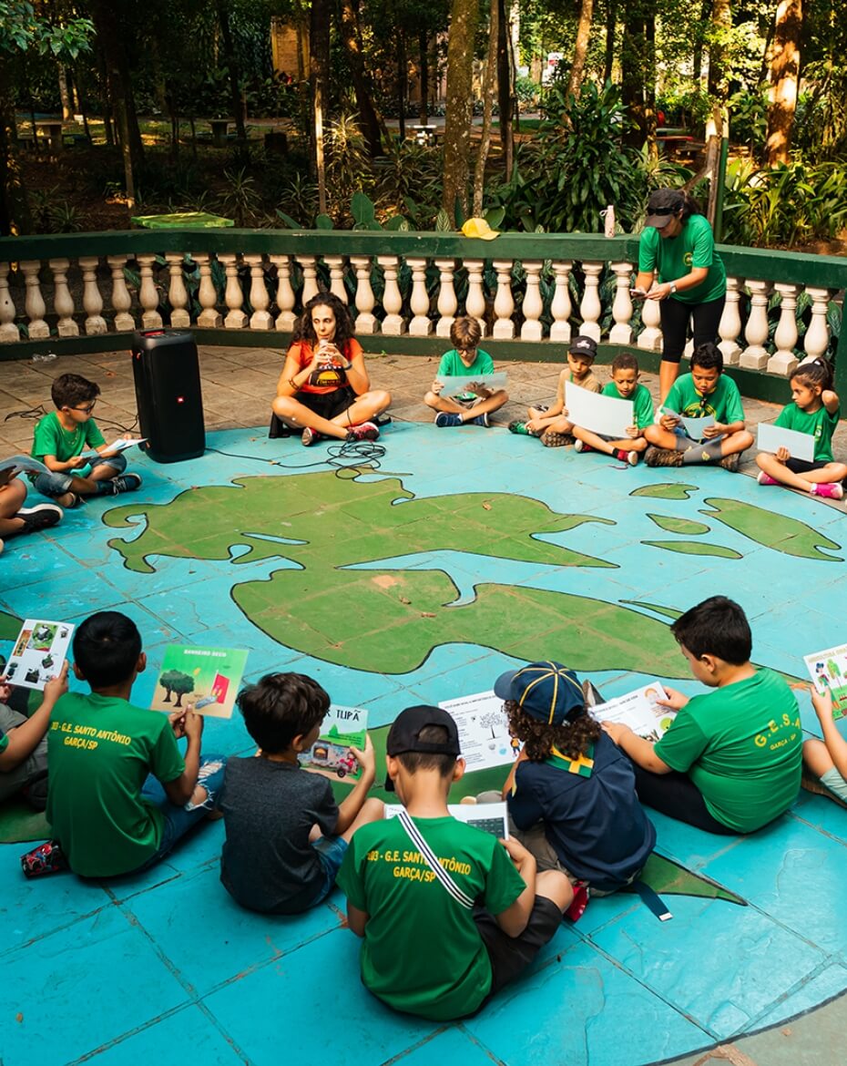 Oficina educativa realizada ao ar livre com um grupo de crianças sentadas em círculo no chão, que possui a pintura de um mapa mundi, lendo livretos sob a orientação de uma instrutora sentada com um microfone.