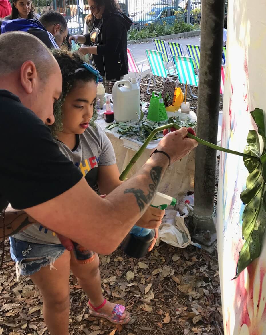 Homem tatuado auxiliando uma menina de cabelos cacheados a manusear um longo caule verde e um frasco de borrifador, com uma mesa repleta de mudas, folhas e insumos ao fundo, em um ambiente de praça com cadeiras de praia listradas.